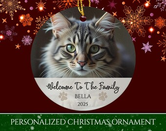 May include: Circular Christmas ornament with a cat's portrait, green eyes, and long whiskers. The ornament has the text "Welcome To The Family", the name "BELLA", and the year "2025". The background is dark red with snowflake designs.