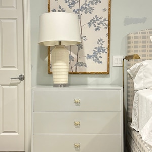 May include: A light gray dresser with three drawers and gold butterfly-shaped drawer pulls. The dresser is in front of a framed print of a blue and white floral design.