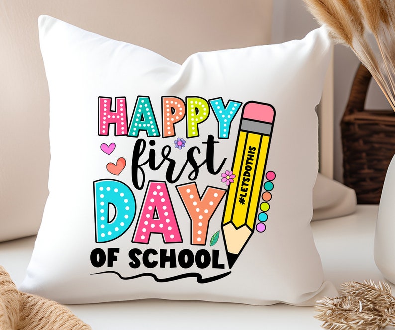 May include: White square pillow with a colorful "Happy First Day of School" design. The text is in various colors and fonts, with a large yellow pencil graphic. The pencil has the text "#LETSDOTHIS" on it.