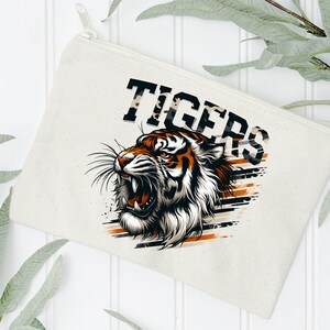 K&ouml;nnte beinhalten: Eine wei&szlig;e Canvas-Rei&szlig;verschlusstasche mit einer Grafik eines br&uuml;llenden Tigers mit orangefarbenen und schwarzen Streifen. Der Text "TIGERS" ist in Schwarz &uuml;ber dem Tiger gedruckt.