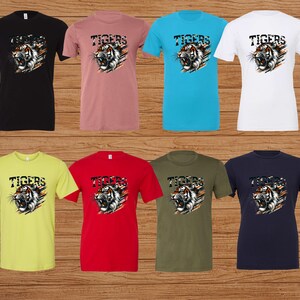 K&ouml;nnte beinhalten: Acht T-Shirts in verschiedenen Farben mit einem Aufdruck eines Tigerkopfes und dem Wort "TIGERS" dar&uuml;ber.