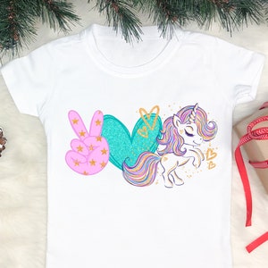 May include: White t-shirt with a pink peace sign, a teal heart, and a rainbow unicorn with gold glitter accents. The text "Peace Love Unicorn" is printed on the shirt.