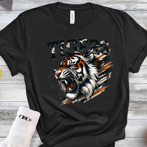 K&ouml;nnte beinhalten: Schwarzes T-Shirt mit einem Tigerkopf-Motiv und dem Wort "TIGERS" in einem Tarnmuster dar&uuml;ber.