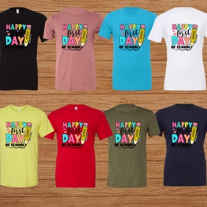 May include: Assortment of t-shirts in various colors, including black, pink, blue, and white. Each shirt features a colorful graphic with the text "Happy First Day of School" and a pencil illustration. The shirts are displayed on a wooden background.