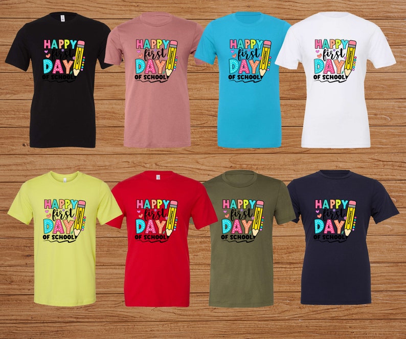May include: Assortment of t-shirts in various colors, including black, pink, blue, and white. Each shirt features a colorful graphic with the text "Happy First Day of School" and a pencil illustration. The shirts are displayed on a wooden background.