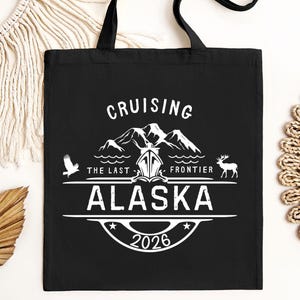 A black tote bag with white text and graphics, including a mountain silhouette, a bird, and the text "CRUISING THE LAST FRONTIER ALASKA 2026".