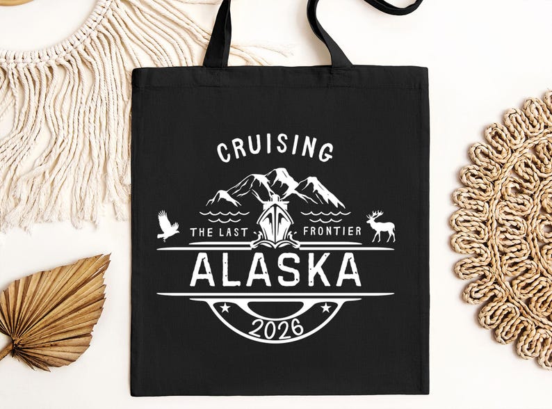 A black tote bag with white text and graphics, including a mountain silhouette, a bird, and the text "CRUISING THE LAST FRONTIER ALASKA 2026".