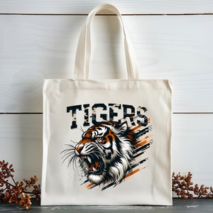 K&ouml;nnte beinhalten: Eine wei&szlig;e Canvas-Tragetasche mit einem Tiger-Motiv und dem Wort "TIGERS" in Schwarz gedruckt. Der Tiger ist orange, schwarz und wei&szlig; mit einem wilden Ausdruck.