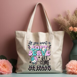 Pode incluir: Uma bolsa tote de lona branca com um gr&aacute;fico rosa, azul e verde que diz "Se voc&ecirc; quer um soft serve, pegue um sorvete". O gr&aacute;fico tamb&eacute;m inclui uma bola de voleibol com um design rosa e azul.
