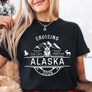 A woman wearing a black t-shirt with a graphic design and text, holding a mug with a Santa Claus design, and standing in front of a Christmas tree.