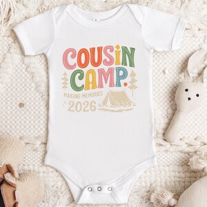 May include: White baby onesie with the text "COUSIN CAMP" in colorful letters. The design includes a tent, trees, and the words "MAKING MEMORIES 2026". The onesie is surrounded by a soft, textured blanket and a stuffed bunny.