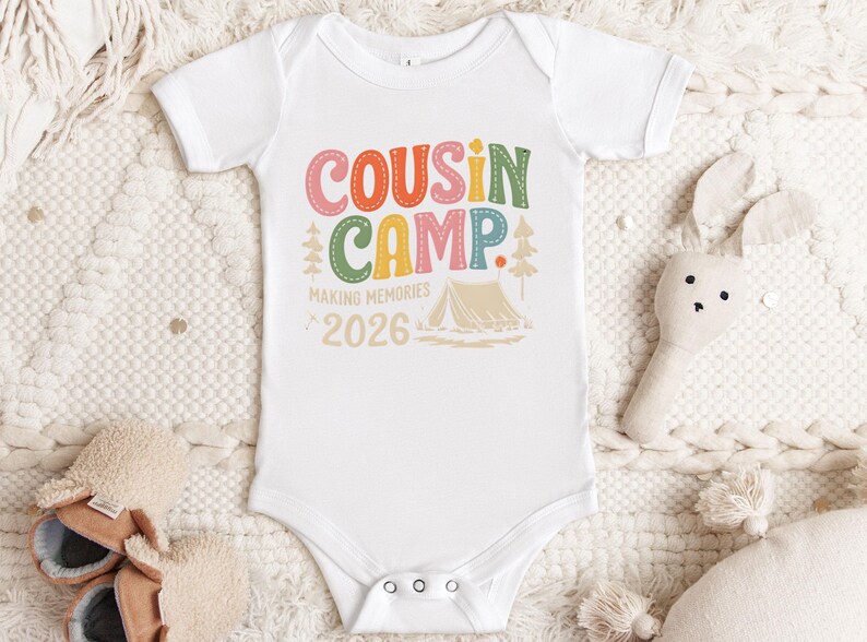 May include: White baby onesie with the text "COUSIN CAMP" in colorful letters. The design includes a tent, trees, and the words "MAKING MEMORIES 2026". The onesie is surrounded by a soft, textured blanket and a stuffed bunny.