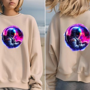 Pode incluir: Camisola bege com um gr&aacute;fico vibrante de um astronauta numa cena espacial. O astronauta est&aacute; dentro de uma moldura circular com um fundo de nebulosa colorida. O design &eacute; repetido na frente e atr&aacute;s da camisola.