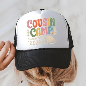 May include: A black and white trucker hat with the words "COUSIN CAMP" in colorful letters. The hat also features the text "MAKING MEMORIES 2026" and a graphic of a tent and trees. The hat is being held by a person.