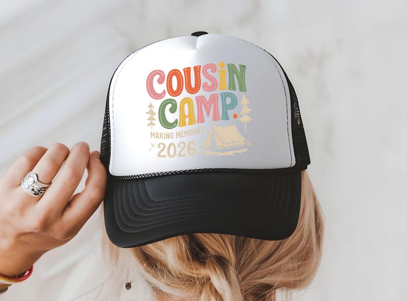 May include: A black and white trucker hat with the words "COUSIN CAMP" in colorful letters. The hat also features the text "MAKING MEMORIES 2026" and a graphic of a tent and trees. The hat is being held by a person.