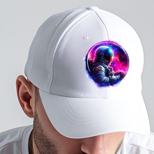 Pode incluir: Bon&eacute; de beisebol branco com um gr&aacute;fico circular de um astronauta no espa&ccedil;o. O astronauta usa um capacete e um traje azuis, em um fundo de nebulosas rosa, roxas e azuis. O bon&eacute; tem um design cl&aacute;ssico de seis pain&eacute;is.
