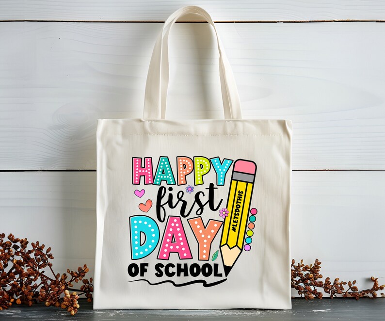 May include: Cream-colored tote bag with the text "HAPPY FIRST DAY OF SCHOOL" in colorful bubble letters. A yellow pencil graphic with the text "#LETSDOTHIS" is on the right side of the bag. The bag has two cream-colored handles.