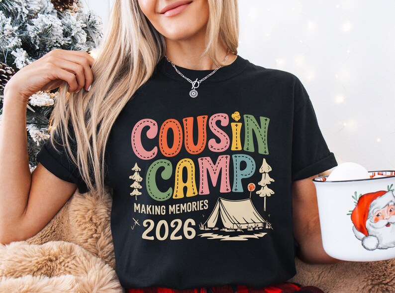 May include: Black t-shirt with the words "COUSIN CAMP" in colorful stitched letters. Below the text is "MAKING MEMORIES 2026" with a tent and trees graphic. A white mug with a Santa Claus design is held in the right hand.