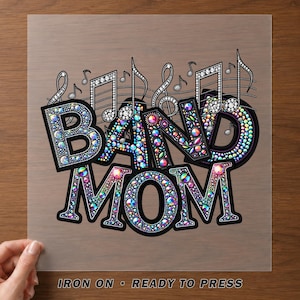 May include: An iron-on transfer sheet featuring the text BAND MOM in bold, black-outlined letters filled with multicolored, iridescent rhinestones. Musical notes and staff lines are positioned above the text, also accented with rhinestones.