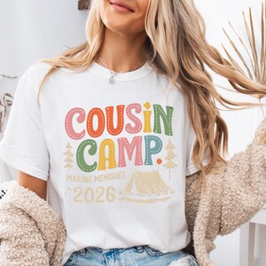 May include: White t-shirt with the words "COUSIN CAMP" in colorful letters. Below the text are the words "MAKING MEMORIES" and the year "2026". A tent and trees are also featured in the design. The shirt is a casual wear item.