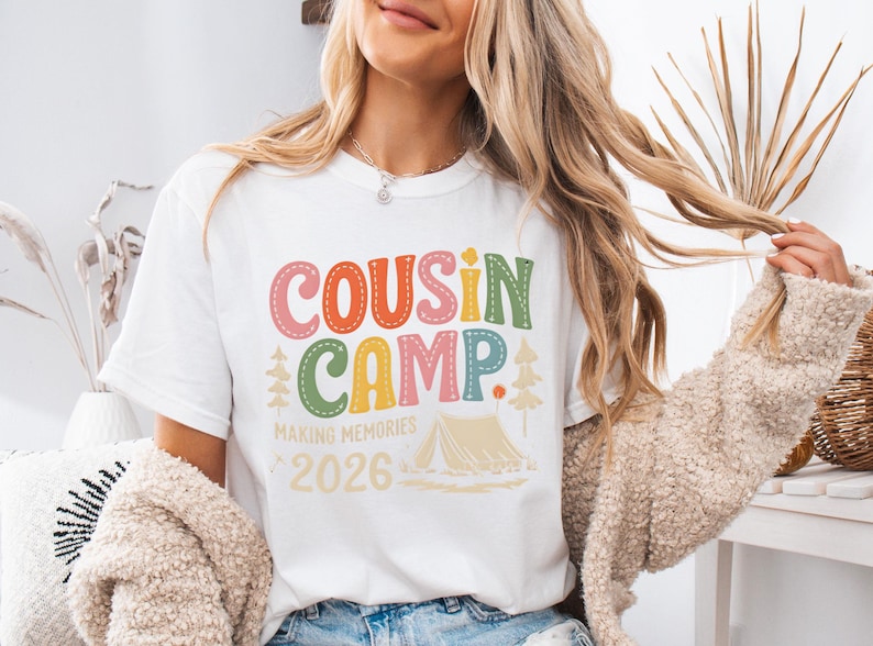 May include: White t-shirt with the words "COUSIN CAMP" in colorful letters. Below the text are the words "MAKING MEMORIES" and the year "2026". A tent and trees are also featured in the design. The shirt is a casual wear item.