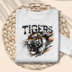 K&ouml;nnte beinhalten: Wei&szlig;er Pullover mit einem Aufdruck eines Tigers mit orangefarbenen und schwarzen Streifen und dem Wort "TIGERS" in schwarzen Buchstaben mit einem Tarnmuster.