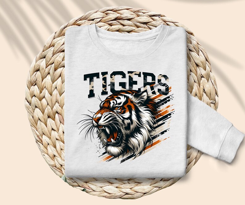 K&ouml;nnte beinhalten: Wei&szlig;er Pullover mit einem Aufdruck eines Tigers mit orangefarbenen und schwarzen Streifen und dem Wort "TIGERS" in schwarzen Buchstaben mit einem Tarnmuster.