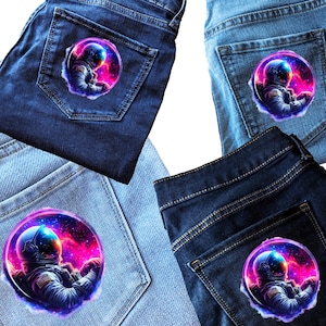 Pode incluir: Quatro pares de cal&ccedil;as de ganga com um patch de astronauta colorido no bolso de tr&aacute;s. O astronauta &eacute; retratado numa cena espacial com um capacete azul e um fundo de nebulosa rosa e roxa vibrante.