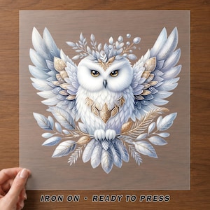 May include: An iron-on transfer featuring a white owl with spread wings, accented by gold and silver leaf patterns and foliage. The owl has amber eyes and is surrounded by decorative feathers and leaves. The text reads IRON ON • READY TO PRESS.