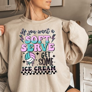 Pode incluir: Uma camisa de moletom bege com uma estampa gr&aacute;fica que diz "Se voc&ecirc; quer um sorvete mole, pegue um sorvete". O gr&aacute;fico inclui uma bola de v&ocirc;lei, raios, estrelas e um padr&atilde;o xadrez.