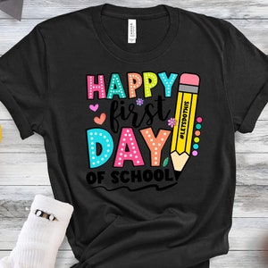 May include: Black t-shirt with a colorful graphic that says "Happy First Day of School." The design includes a large yellow pencil with the text "#LetsDoThis." The text is in various colors, including pink, blue, and green.