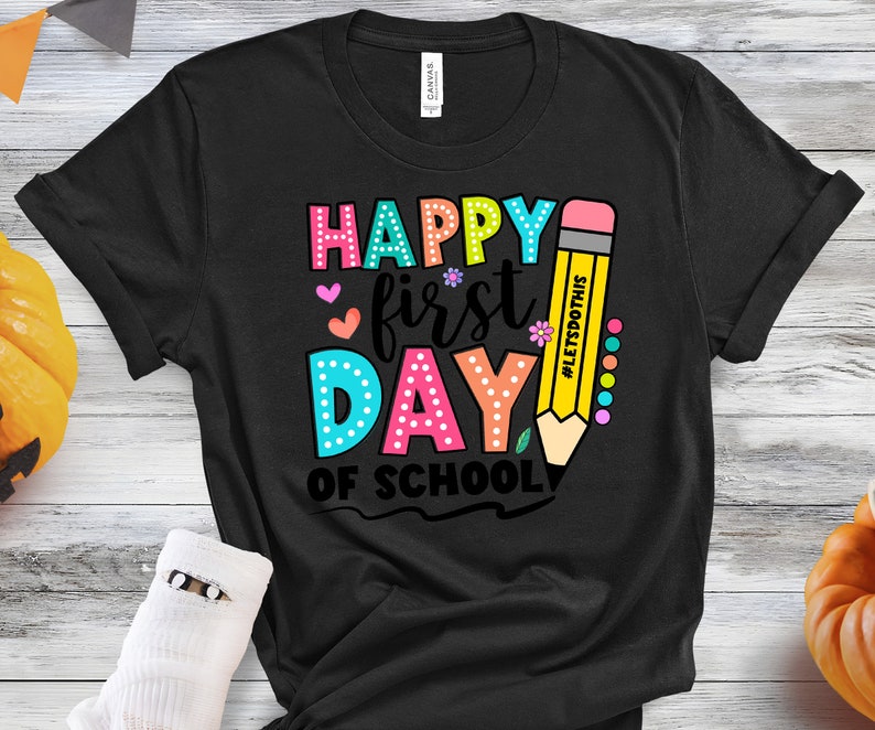 May include: Black t-shirt with a colorful graphic that says "Happy First Day of School." The design includes a large yellow pencil with the text "#LetsDoThis." The text is in various colors, including pink, blue, and green.