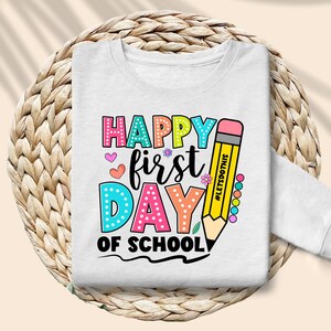 May include: White long-sleeved sweatshirt with a colorful graphic that says "Happy First Day of School." The design includes a large yellow pencil and various hearts and flowers. The text "#LETSDOTHIS" is on the pencil.