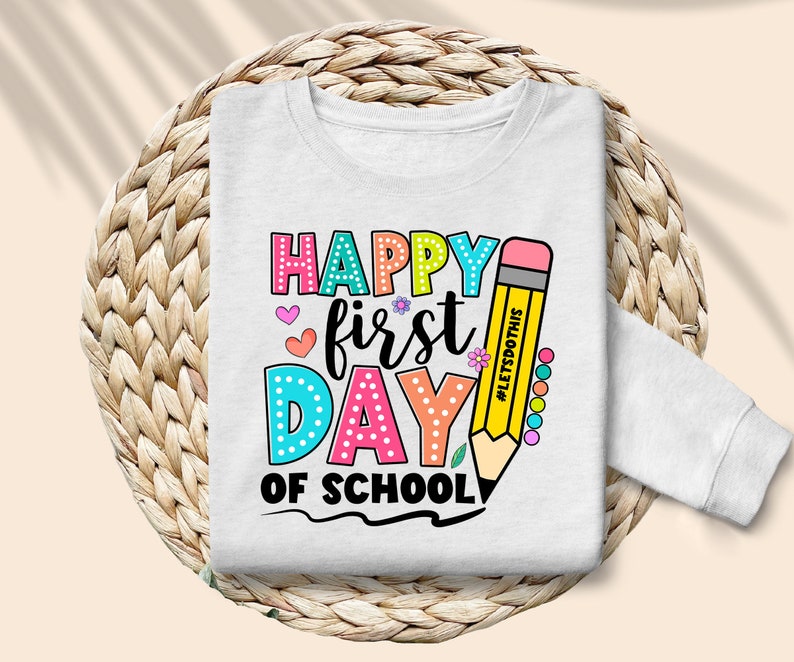 May include: White long-sleeved sweatshirt with a colorful graphic that says "Happy First Day of School." The design includes a large yellow pencil and various hearts and flowers. The text "#LETSDOTHIS" is on the pencil.