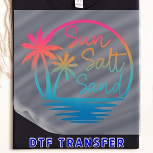 May include: Black t-shirt with a DTF transfer featuring a colorful design. The design includes a circle with the words "Sun, Salt, Sand" and palm trees. The text "DTF TRANSFER" is at the bottom.