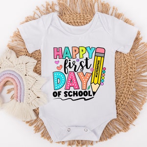 May include: White baby onesie with a colorful "Happy First Day of School" graphic. The design includes a large yellow pencil, hearts, and flowers. The text "#LetsDoThis" is also included.