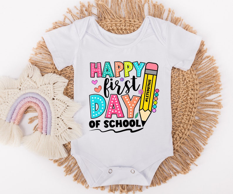 May include: White baby onesie with a colorful "Happy First Day of School" graphic. The design includes a large yellow pencil, hearts, and flowers. The text "#LetsDoThis" is also included.