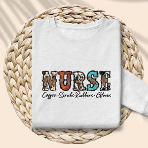 May include: White sweatshirt with the word "NURSE" in colorful patterned letters. The text "Coffee. Scrubs. Rubbers. Gloves" is printed below the word "NURSE".