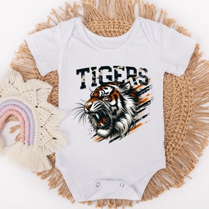K&ouml;nnte beinhalten: Wei&szlig;er Baby-Strampler mit einem Tiger-Motiv und dem Wort "TIGERS" in schwarzen und orangen Buchstaben. Der Tiger ist orange und schwarz vor wei&szlig;em Hintergrund.