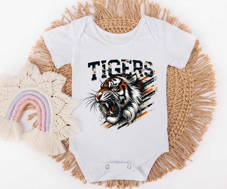 K&ouml;nnte beinhalten: Wei&szlig;er Baby-Strampler mit einem Tiger-Motiv und dem Wort "TIGERS" in schwarzen und orangen Buchstaben. Der Tiger ist orange und schwarz vor wei&szlig;em Hintergrund.