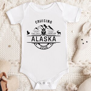 A white onesie with a graphic design and text, laid out on a white background with a stuffed animal and a pair of shoes.