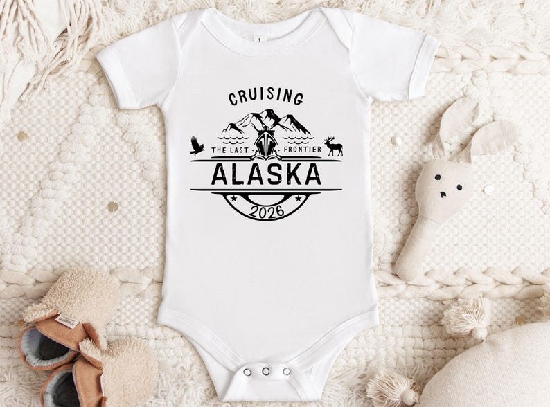 A white onesie with a graphic design and text, laid out on a white background with a stuffed animal and a pair of shoes.