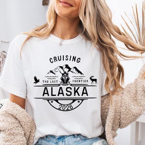 A woman wearing a white t-shirt with a graphic design that says "Cruising the Last Frontier Alaska 2026".