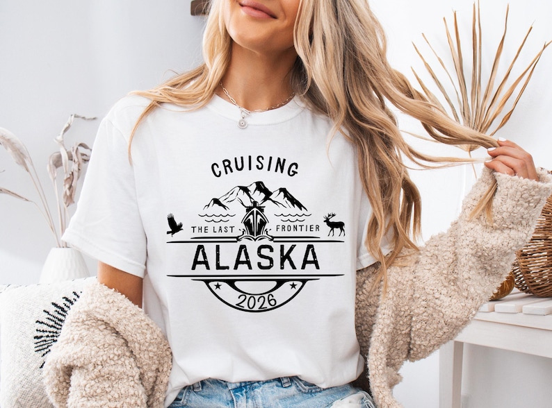A woman wearing a white t-shirt with a graphic design that says "Cruising the Last Frontier Alaska 2026".