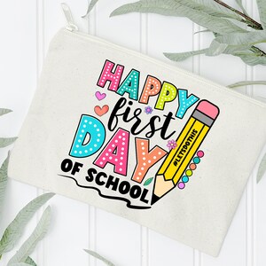 May include: A beige canvas pencil case with a zipper closure. The case features the text "HAPPY first DAY of SCHOOL" in colorful bubble letters, along with a yellow pencil illustration. The pencil has the text "#LETSDOTHS" on it.