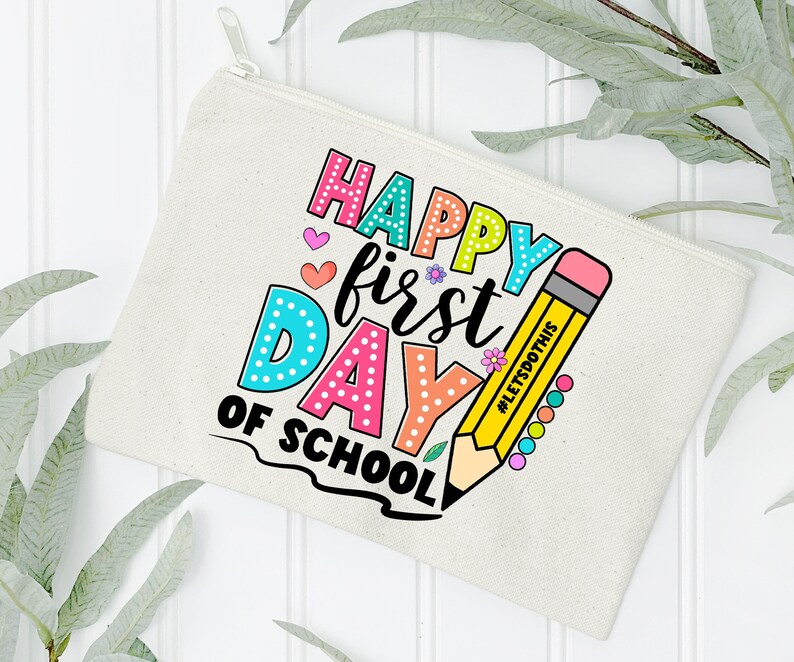 May include: A beige canvas pencil case with a zipper closure. The case features the text "HAPPY first DAY of SCHOOL" in colorful bubble letters, along with a yellow pencil illustration. The pencil has the text "#LETSDOTHS" on it.