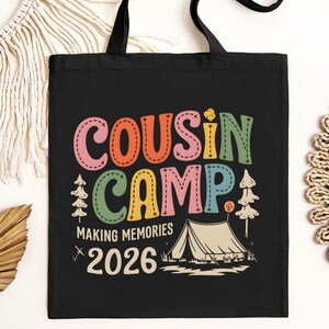 May include: Black tote bag with the words "COUSIN CAMP" in colorful, stitched-style letters. Below, it reads "MAKING MEMORIES 2026" with a tent graphic. The bag has black handles.