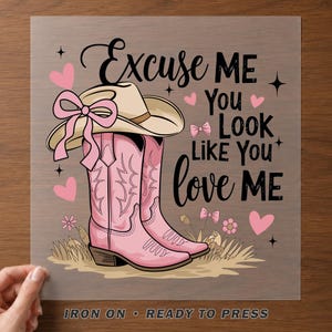 May include: A graphic transfer featuring a beige cowboy hat with a pink bow, pink cowboy boots, and small pink hearts and flowers. The text reads: Excuse ME You LOOK LIKE YOU love ME. The design is on a clear sheet labeled IRON ON READY TO PRESS.