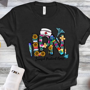 Peut inclure: T-shirt noir avec un graphique color&eacute; des lettres LPN, repr&eacute;sentant une infirmi&egrave;re auxiliaire dipl&ocirc;m&eacute;e, avec des symboles m&eacute;dicaux et des tournesols.