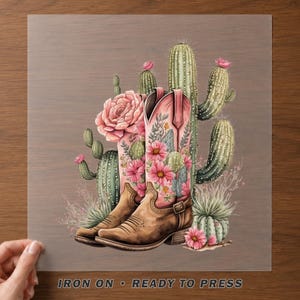 May include: An iron-on transfer featuring a pair of light brown cowboy boots decorated with pink floral patterns, surrounded by various green cacti and succulents, with the text IRON ON READY TO PRESS printed at the bottom of the design.
